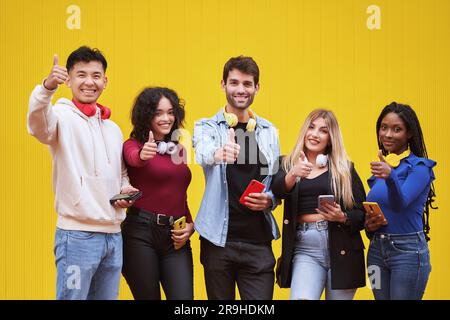Multi-ethnic people showing thumbs up to camera and smiling while standing together against a yellow wall. Friendship, positivity and technology concept. Stock Photo