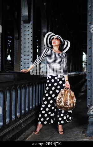 A stylish, glamourous mature woman surrounded by iron columns under Pont Alexandre III Paris, wearing stripes and polka dots, black and white & gold Stock Photo