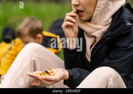 A woman holds various dried fruits and nuts in her hand. Sits on the green grass in the forest. Snack during the hike, walk. Healthy vegetarian food. Stock Photo