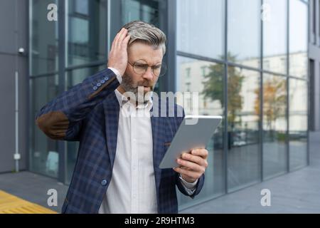 Mature gray haired boss businessman received message with bad news, man shocked by bankruptcy and upset reading message from outside office building, holding tablet. Stock Photo