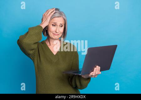 Portrait of unhappy dissatisfied woman wear khaki sweater look at laptop hand on head got critical error isolated on blue color background Stock Photo