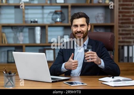 Portrait of successful mature businessman inside office at workplace, senior experienced man smiling and looking at camera, using computer at work, drinking water and showing thumbs up encouraging. Stock Photo