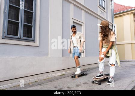 Tourists couple skateboarders riding skates. Happy young travelers longboarding together on sunset on pavement. Stylish man and woman in trendy outfit Stock Photo