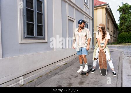 Tourists couple skateboarders riding skates. Happy young travelers longboarding together on sunset on pavement. Stylish man and woman in trendy outfit Stock Photo