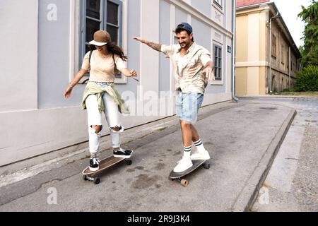 Tourists couple skateboarders riding skates. Happy young travelers longboarding together on sunset on pavement. Stylish man and woman in trendy outfit Stock Photo