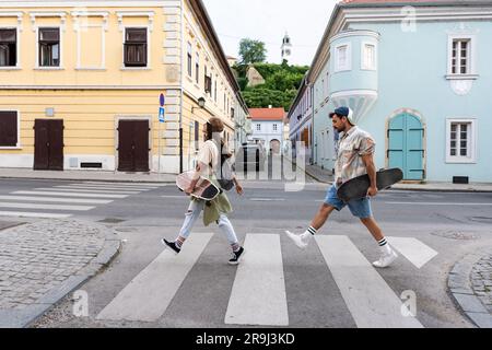 Tourists couple skateboarders riding skates. Happy young travelers longboarding together on sunset on pavement. Stylish man and woman in trendy outfit Stock Photo