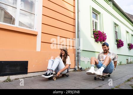 Tourists couple skateboarders riding skates. Happy young travelers longboarding together on sunset on pavement. Stylish man and woman in trendy outfit Stock Photo