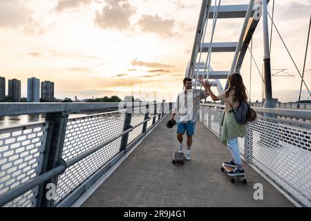 Tourists couple skateboarders riding skates. Happy young travelers longboarding together on sunset on pavement. Stylish man and woman in trendy outfit Stock Photo