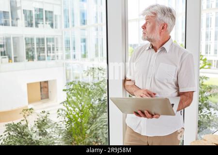 Thoughtful senior businessman with laptop looking out of window Stock Photo