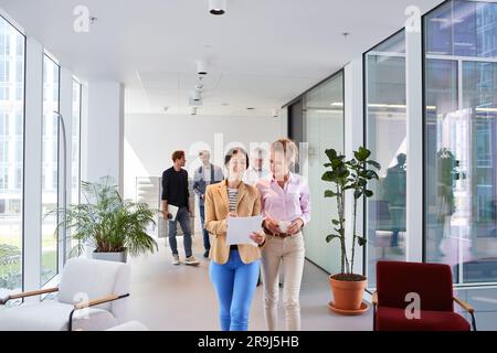 Smiling businesswomen discussing over documents while walking in corridor Stock Photo