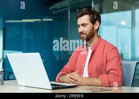 Mature adult businessman working on laptop inside office, programmer smiling contentedly reading and checking software code, man in red shirt. Stock Photo