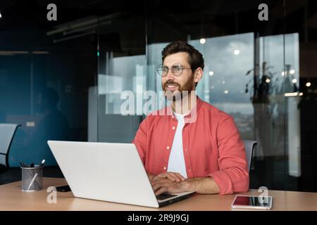 Smiling dreamy businessman inside office at workplace, experienced and successful programmer engineer smiling and looking out window, satisfied with results of achievement at work. Stock Photo