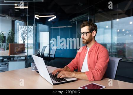 Thinking and serious man working with laptop inside office, focused programmer coding new software. Stock Photo