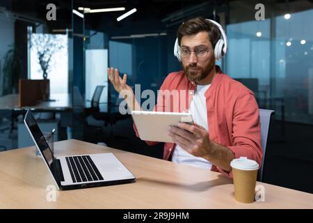 Man with headphones and tablet computer at workplace disappointed with game result, businessman programmer watching online sports match and cheering. Stock Photo