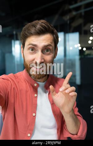 A businessman looks into the camera of the phone, pov. man inside office talking with friends smiling using app on smartphone for video call. Vertical shot Stock Photo