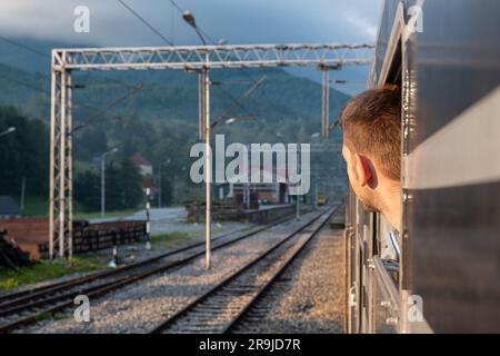 Young man looking out the train window while train arriving to the station at sunrise. Male traveling by railway through Balkans. Stock Photo