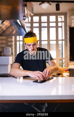 Focused young male chef with yellow headband looking down while arranging sushi roll on dark stone plate at sushi bar Stock Photo