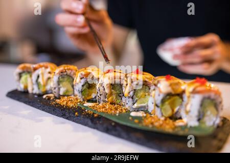 Closeup of anonymous person with chopsticks serving tobiko on uramaki sushi rolls with avocado eel and rice on wooden tray against blurred interior Stock Photo