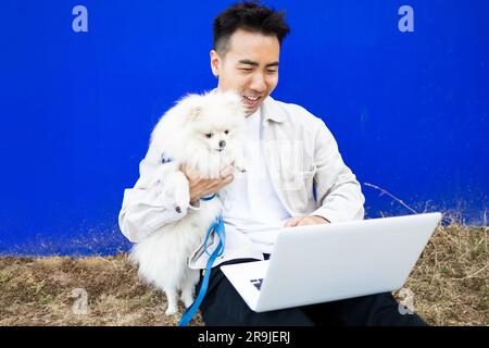 Happy Asian male dog owner in casual wear with cute Pomeranian Spitz and computer video calling while relaxing on weekend Stock Photo