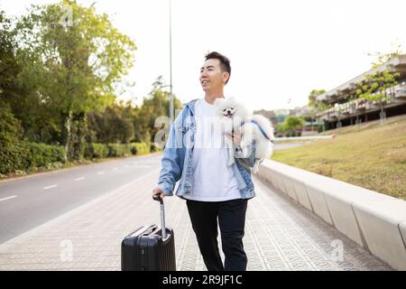 Happy Asian male tourist in casual wear with suitcase and cute Pomeranian Spitz dog smiling and strolling on street Stock Photo