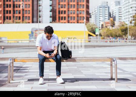 Serious young Hispanic male in sunglasses sitting on wooden bench with backpack while looking at screen and browsing mobile phone in daylight against Stock Photo