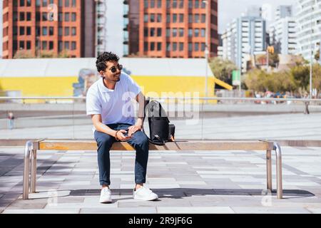 Positive young Hispanic male in sunglasses smiling and sitting on wooden bench with backpack while looking away in daylight against blurred buildings Stock Photo