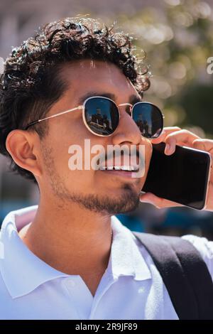 Ethnic bearded guy in trendy sunglasses smiling and looking away while speaking on smartphone in sunlight Stock Photo