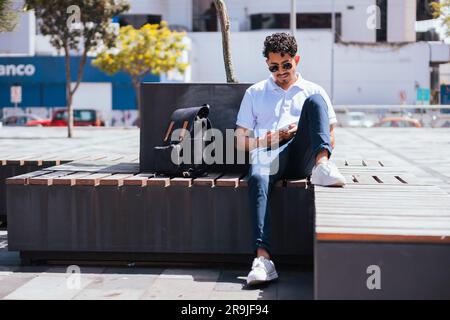 Full body of Hispanic male in sunglasses sitting on bench and browsing mobile phone while resting on sunny day in city Stock Photo