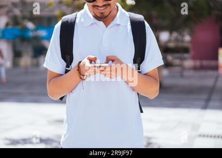 Anonymous young male in sunglasses with backpack looking at screen of mobile phone and browsing while standing on city street near modern Stock Photo