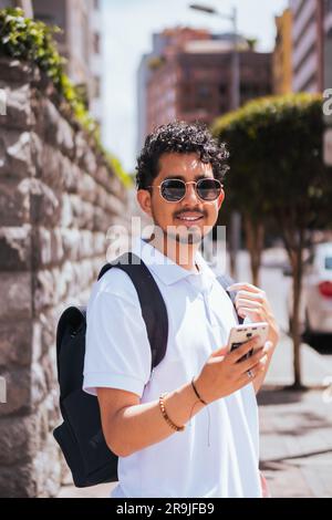 Happy Hispanic male in sunglasses looking at camera while standing on street near stone wall and using cellphone in daylight Stock Photo