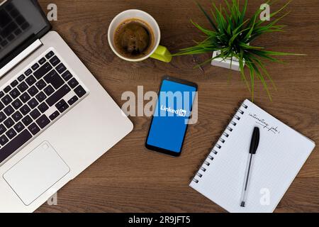 Linkedin app on phone screen. Workspace top view. Table desk with laptop, mobile phone, coffee, notepad, pen and plant. Job search and jobs website.RO Stock Photo