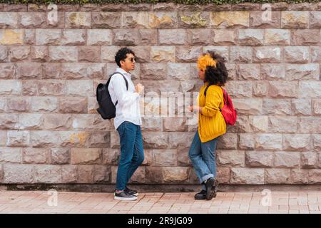 Side view of young friends in casual clothes with backpacks facing each other while standing on paved street against stone wall talking and looking at Stock Photo