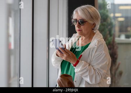 Focused aged lady with short gray hair in sunglasses surfing internet on smartphone while standing near window Stock Photo