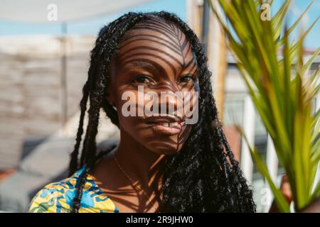 Portrait of smiling young African American female with long dreadlocks looking at camera while standing near palm leaf on sunny day Stock Photo
