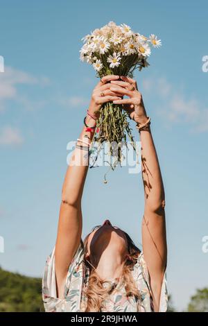 Unrecognizable female with fresh blooming bouquet of white wild daisy flowers on summer vacation on green field against blue sky Stock Photo