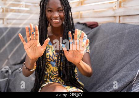 Smiling young African American female in casual clothes with long dreadlocks showing palms with blue paint looking at camera Stock Photo