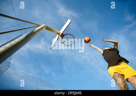 Low angle of unrecognizable female sportswoman shooting ball in hoop on billboard against blue cloudy sky Stock Photo