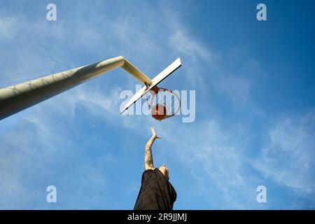 Low angle of unrecognizable masculine sportswoman shooting ball in hoop on billboard against blue cloudy sky Stock Photo