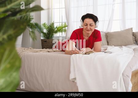 Side view of focused mature female in red dress lying on soft bed and writing information in notebook while enjoying music in headphones Stock Photo