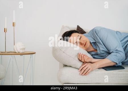 Calm middle aged female in blue shirt lying on comfortable sofa and napping in light living room Stock Photo