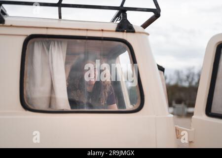 Through glass view of positive young female smiling and looking away while sitting near window with curtain in parked van with opened door Stock Photo