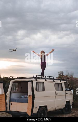 Back view of young traveler in trainers standing on parked van top with open arms Stock Photo
