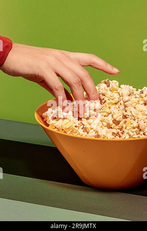 Crop anonymous female with hand picking delicious popcorn heap in yellow bowl against green background Stock Photo