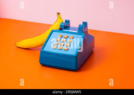 From above of old fashioned plastic toy table phone with fresh ripe banana placed on plain orange surface over bright pink background in studio Stock Photo