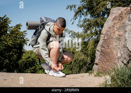 Young short haired female traveler with backpack and fitness mat tying shoelace on sneaker and standing near stones on path with nature at background, Stock Photo