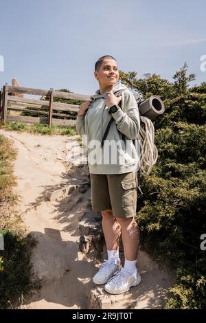 Smiling and short haired female hiker with backpack, fitness mat and climbing rope standing and looking away on hill with sky at background, hiker fin Stock Photo