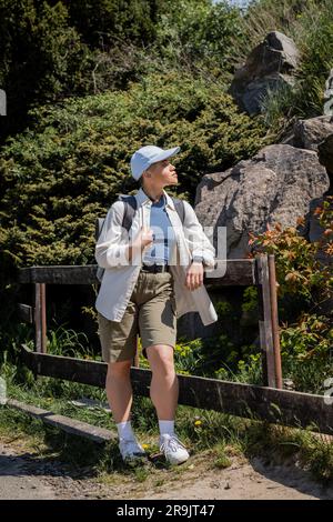 Side view of young short haired female traveler in casual clothes and baseball cap with backpack looking away while standing near fence outdoors, Tran Stock Photo
