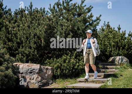 Young female hiker in casual clothes and baseball cap holding backpack while walking down on stairs with nature and blue sky at background, curious hi Stock Photo