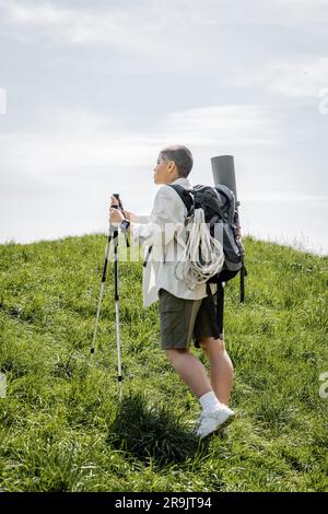 Side view of young short haired female traveler with backpack and climbing rope walking with trekking poles on hill with grass at background, independ Stock Photo