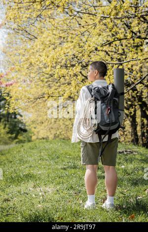 Side view of young and short haired female tourist with backpack, climbing rope and fitness mat standing on glass with landscape at background, indepe Stock Photo